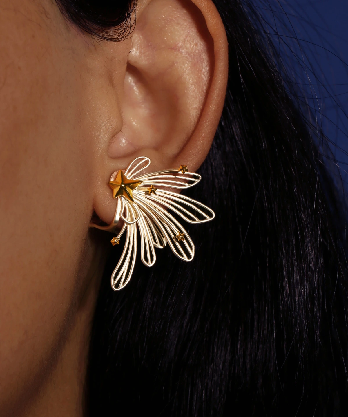 A close-up of a woman's ear showcases the Star Trail 925 Sterling Silver Star Stud Earrings, featuring delicate silver loops and gold star accents radiating from a central gold star. The design evokes the motion of a shooting star.