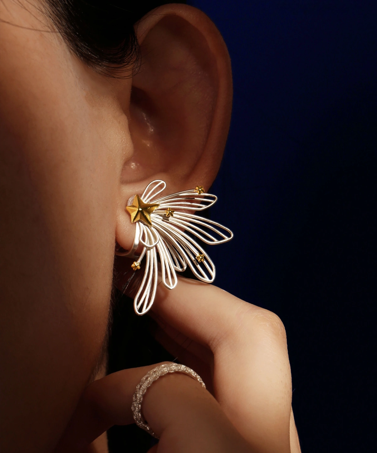 A close-up of a woman's ear showcases the Star Trail 925 Sterling Silver Star Stud Earrings, featuring delicate silver loops and gold star accents radiating from a central gold star. The design evokes the motion of a shooting star.