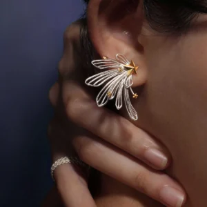 A close-up of a woman's ear showcases the Star Trail 925 Sterling Silver Star Stud Earrings, featuring delicate silver loops and gold star accents radiating from a central gold star. The design evokes the motion of a shooting star.