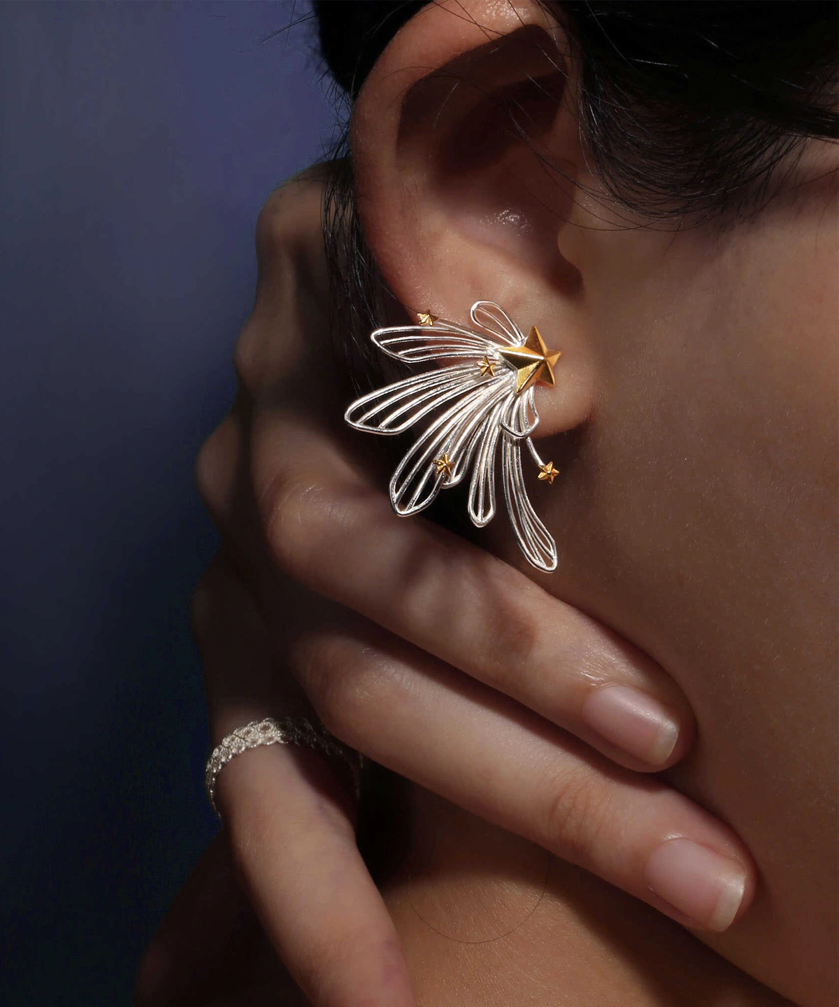 A close-up of a woman's ear showcases the Star Trail 925 Sterling Silver Star Stud Earrings, featuring delicate silver loops and gold star accents radiating from a central gold star. The design evokes the motion of a shooting star.