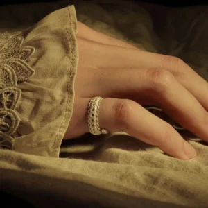 A close-up of a delicate lace ring in S925 sterling silver, elegantly worn on a hand resting on beige fabric with lace embroidery. The adjustable ring features an intricate vintage lace design, blending classic craftsmanship with contemporary style.
