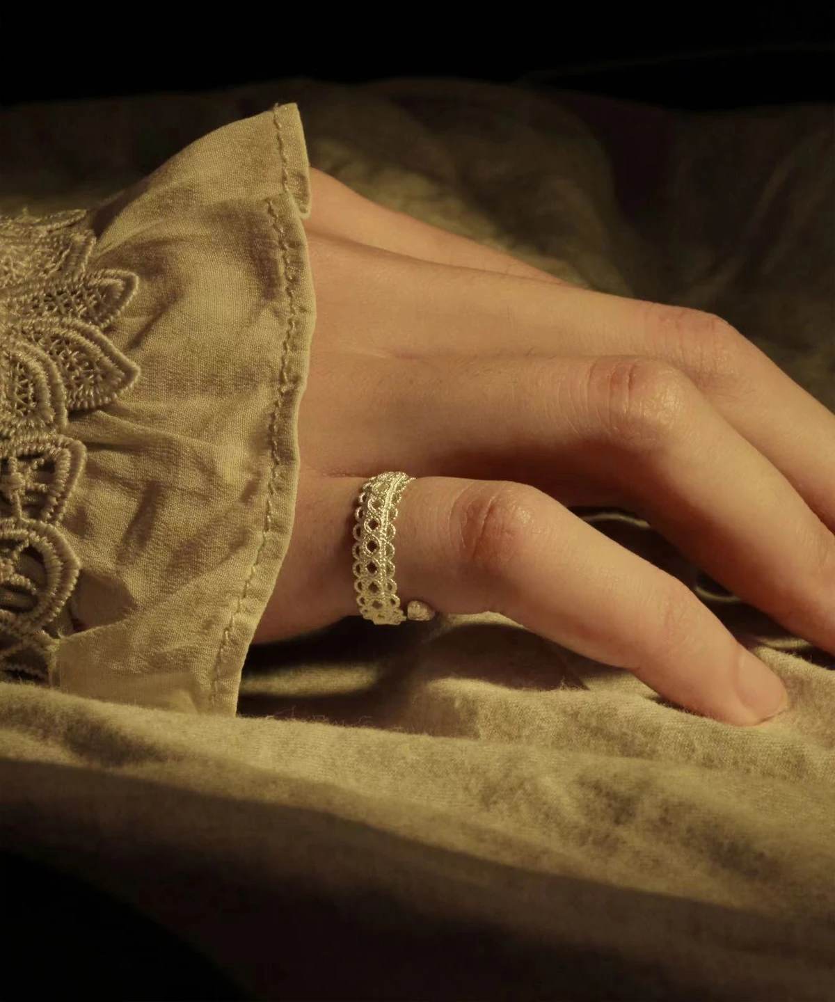 A close-up of a delicate lace ring in S925 sterling silver, elegantly worn on a hand resting on beige fabric with lace embroidery. The adjustable ring features an intricate vintage lace design, blending classic craftsmanship with contemporary style.