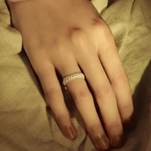 A close-up image of a hand resting on a beige fabric surface, showcasing a Lace Ring on the ring finger. The ring is adjustable and crafted from S925 sterling silver, featuring intricate lace-like patterns that highlight the craftsmanship and elegance of the jewelry piece.