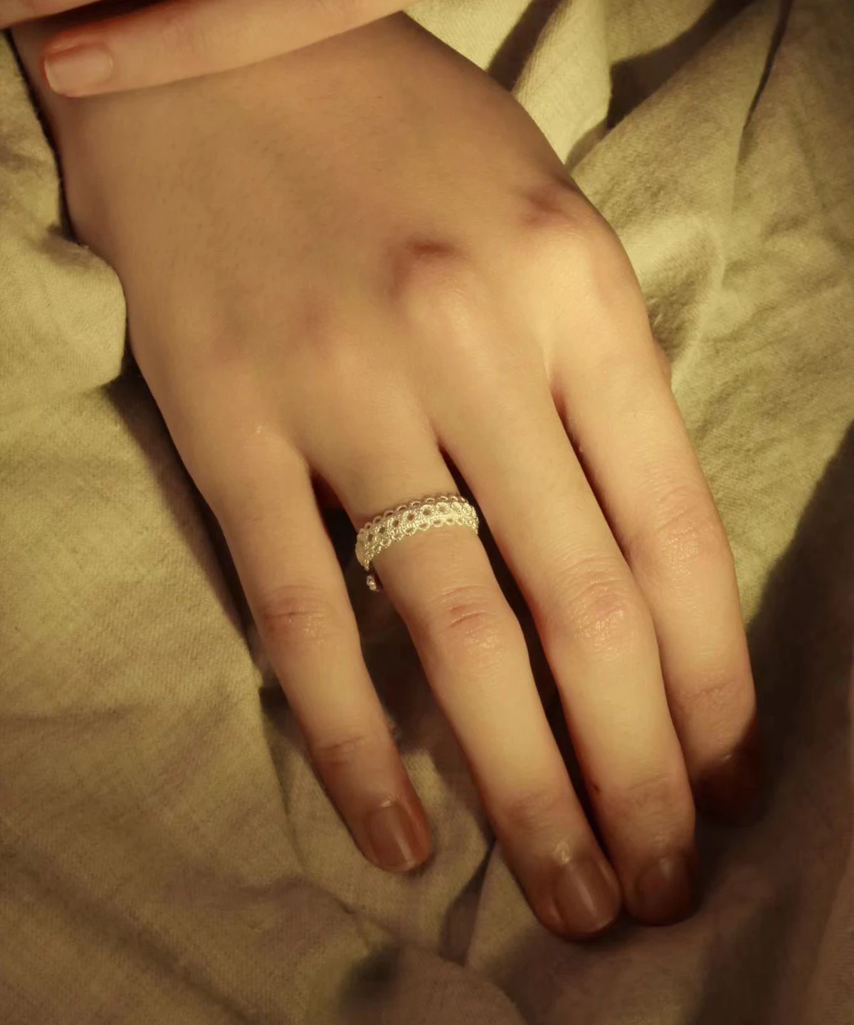 A close-up image of a hand resting on a beige fabric surface, showcasing a Lace Ring on the ring finger. The ring is adjustable and crafted from S925 sterling silver, featuring intricate lace-like patterns that highlight the craftsmanship and elegance of the jewelry piece.