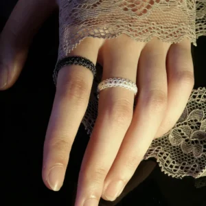 A close-up image of a hand wearing two lace rings, one black and one white, made of S925 sterling silver. The hand is partially covered by delicate lace fabric, highlighting the intricate design and craftsmanship of the rings.