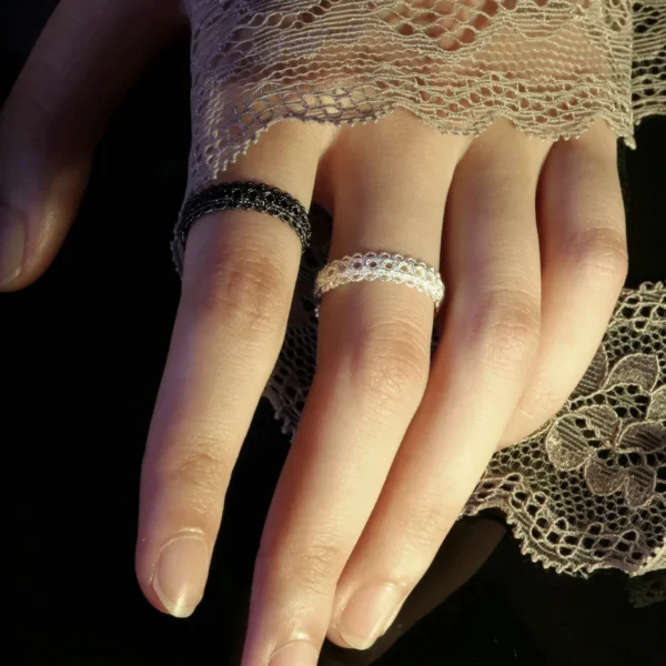 A close-up image of a hand wearing two lace rings, one black and one white, made of S925 sterling silver. The hand is partially covered by delicate lace fabric, highlighting the intricate design and craftsmanship of the rings.