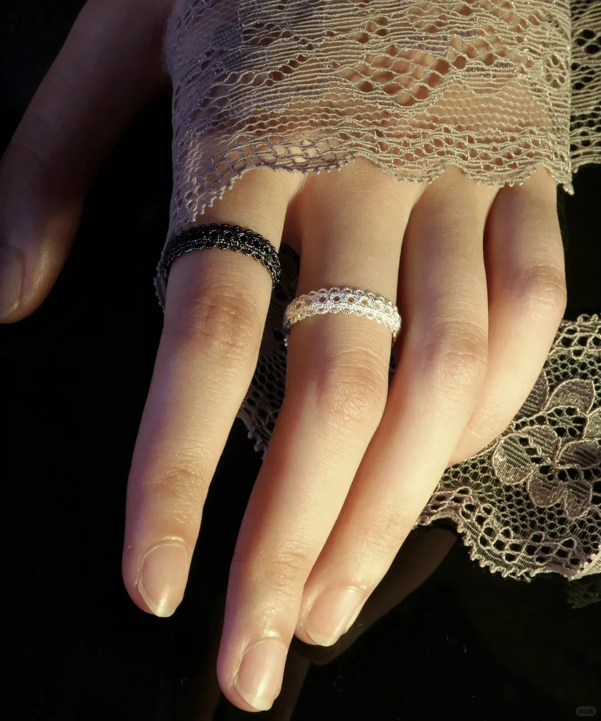 A close-up image of a hand wearing two lace rings, one black and one white, made of S925 sterling silver. The hand is partially covered by delicate lace fabric, highlighting the intricate design and craftsmanship of the rings.
