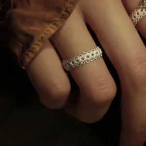 A close-up of a hand adorned with a Lace Ring, crafted from S925 sterling silver. The ring boasts delicate lace-inspired patterns and an adjustable design. The hand, partially draped in a brown fabric sleeve, enhances the vintage aesthetic.