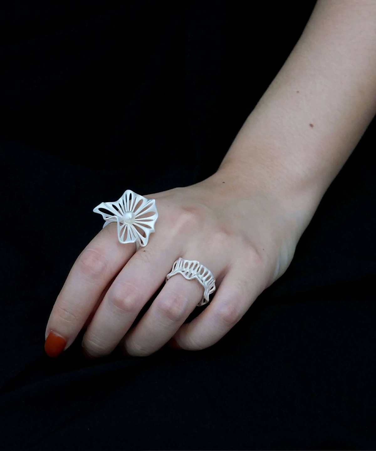 A close-up of a hand wearing a 925 silver adjustable leaf ring with a freshwater pearl on the index finger. The ring features an intricate leaf design, symbolizing elegance and nature-inspired beauty. Another ring with a similar design is worn on the middle finger. The black background enhances the visual appeal.