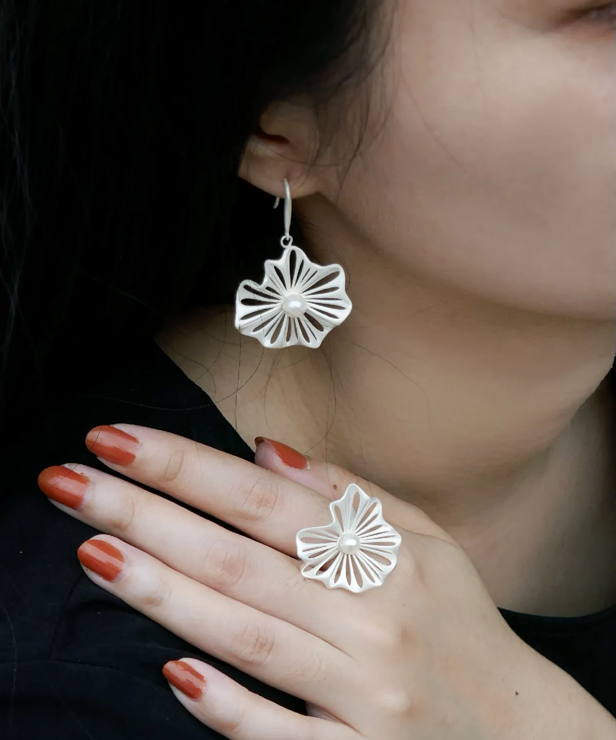 A woman's hand elegantly showcases a 925 silver adjustable leaf ring with a freshwater pearl, accompanied by matching leaf-shaped earrings. The delicate design of the ring features intricate silver detailing resembling leaves, embracing a luminous central pearl. The jewelry complements her reddish-brown nail polish, creating a sophisticated and stylish look.