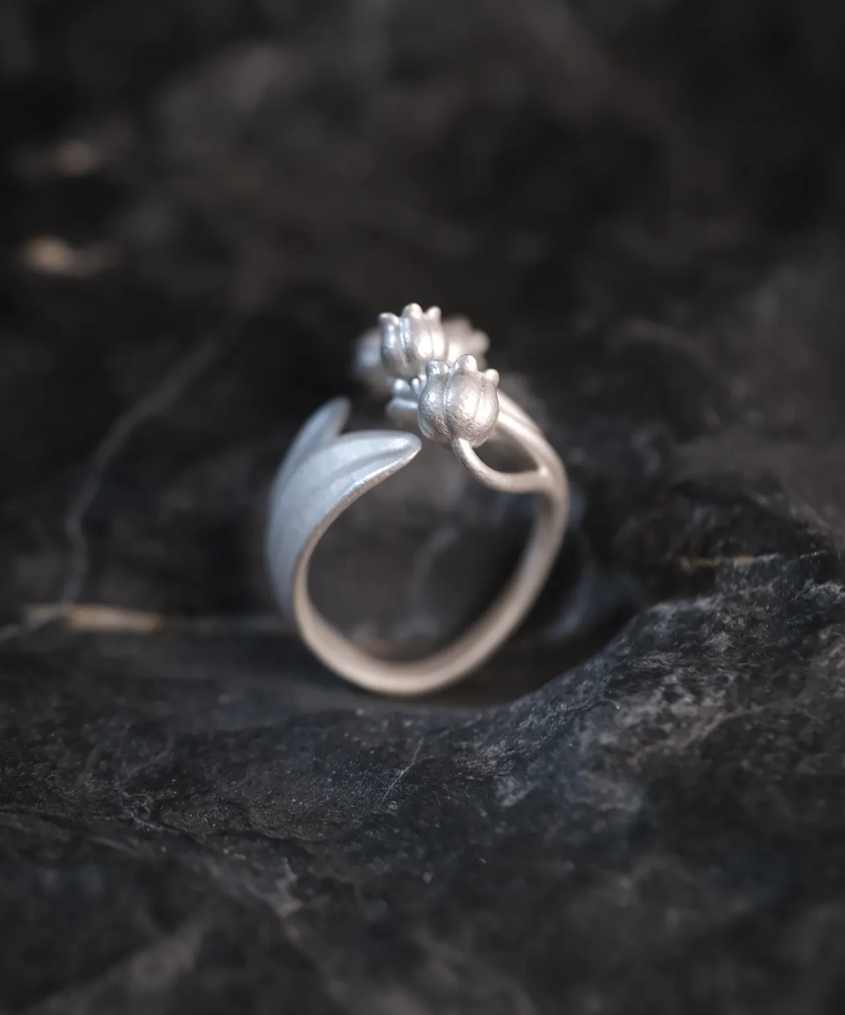 A close-up of the Lily of the Valley Ring, featuring two elegantly sculpted flower buds and delicate leaves, handcrafted from S925 sterling silver. The adjustable silver open ring is set against a dark, textured background, accentuating its intricate detailing.