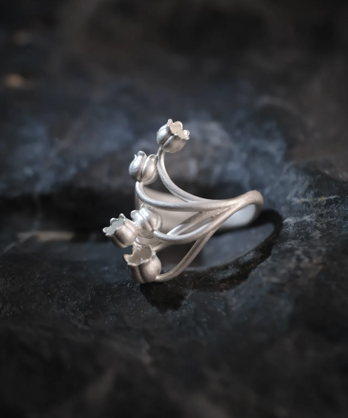 A close-up image of a Lily of the Valley Ring made from S925 sterling silver. The ring features an open design with delicate, handcrafted lily of the valley flowers, elegantly positioned on a dark, textured background.
