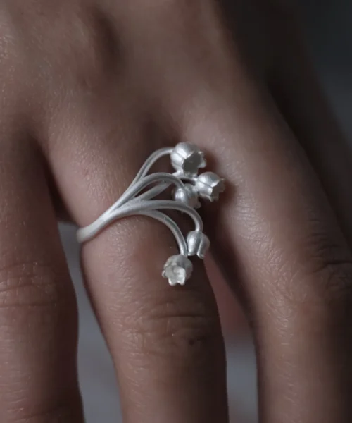 A close-up image of a hand wearing the Lily of the Valley Ring, crafted in S925 sterling silver. The silver open ring features delicate, bell-shaped flowers gracefully extending from the adjustable band.