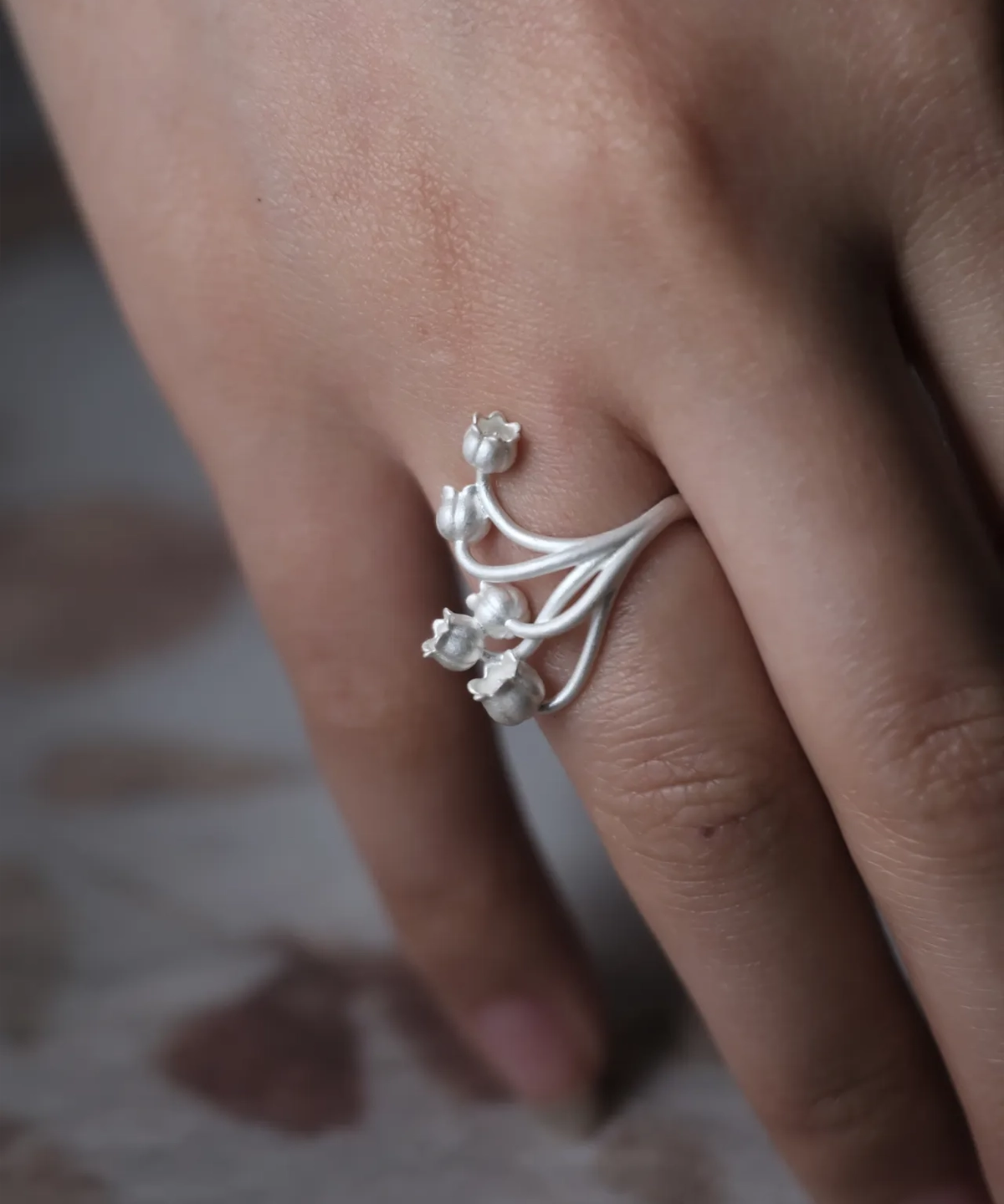 A close-up image of a hand wearing the Lily of the Valley Ring, crafted in S925 sterling silver. The silver open ring features delicate, bell-shaped flowers gracefully extending from the adjustable band.
