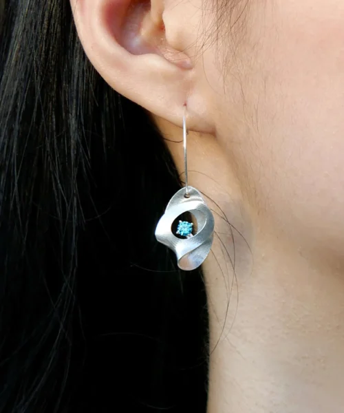Close-up of Lazesoul Nebula Sculptural S925 Silver Drop Earrings worn on the ear, featuring a fluid silver design with a central blue sapphire gemstone — artisan handmade jewelry.