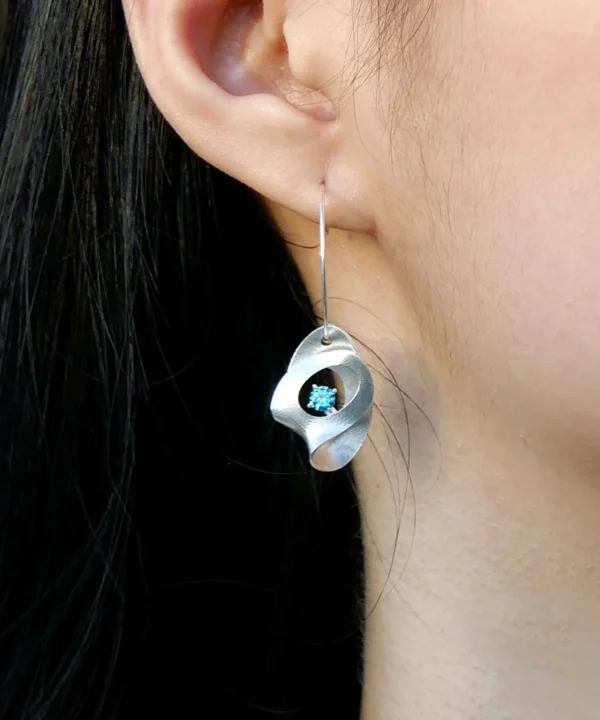 Close-up of Lazesoul Nebula Sculptural S925 Silver Drop Earrings worn on the ear, featuring a fluid silver design with a central blue sapphire gemstone — artisan handmade jewelry.