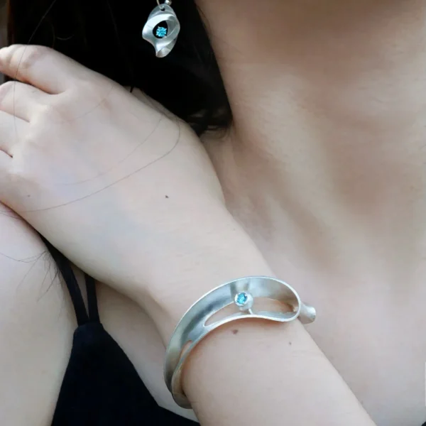 Nebula-Sculptural S925 Sterling Silver Open Bangle 01 A woman's wrist wearing the Nebula Sculptural S925 Sterling Silver Open Bangle with Blue Sapphire—artistic design with fluid curves, complemented by a matching earring, both handmade by Lazesoul.