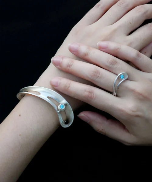 Close-up of a hand adorned with the Lazesoul handmade Nebula S925 sterling silver open bangle featuring a sculptural design and a blue sapphire, paired with a matching ring. Elegant handcrafted silver open bangle jewelry.