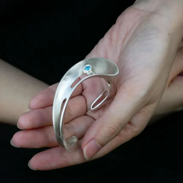 Nebula-Sculptural S925 Sterling Silver Open Bangle 05 A hand holds the Nebula S925 sterling silver open bangle with blue sapphire. This sculptural, handmade art jewelry piece by Lazesoul features a polished finish and openwork design, highlighting the elegance of this silver open bangle.