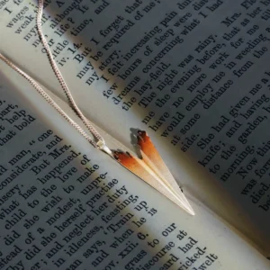 A close-up shot of a Paper Airplane Necklace crafted in S925 Sterling Silver, carefully placed on an open book with elegant printed text. The necklace features a sleek, folded-paper-inspired pendant with a delicate silver chain, reflecting light beautifully. The setting enhances the artistic and handcrafted nature of the jewelry, making it a standout piece from Lazesoul Handmade Art Jewelry.