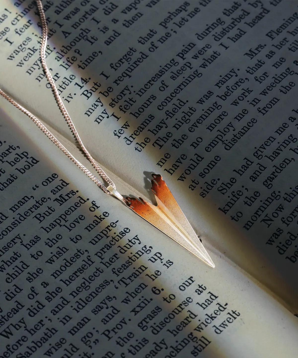 A close-up shot of a Paper Airplane Necklace crafted in S925 Sterling Silver, carefully placed on an open book with elegant printed text. The necklace features a sleek, folded-paper-inspired pendant with a delicate silver chain, reflecting light beautifully. The setting enhances the artistic and handcrafted nature of the jewelry, making it a standout piece from Lazesoul Handmade Art Jewelry.