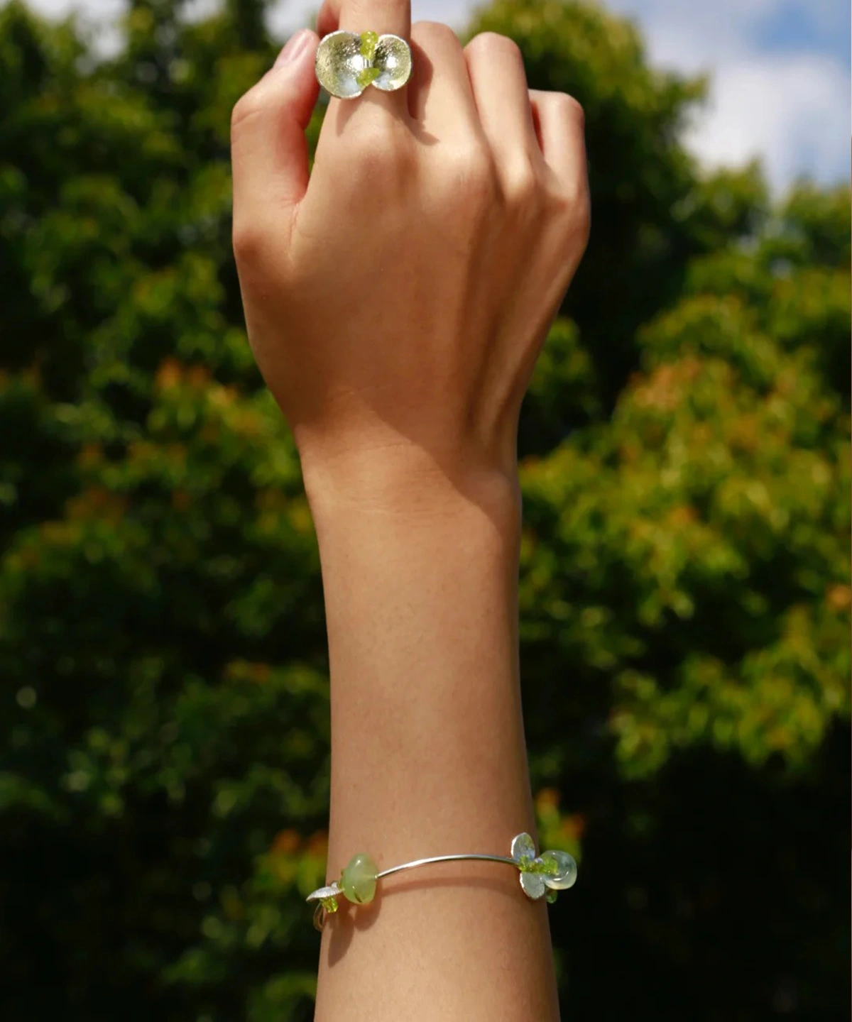 Hand wearing a Lazesoul Prelude Summer peridot bracelet, crafted from S925 sterling silver with natural prehnite and peridot stones, set against a vivid green landscape and blue sky—highlighting the fresh elegance of this handmade jewelry piece.
