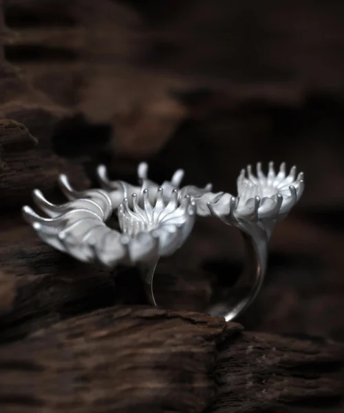 A handcrafted Spiral Ring made of S925 sterling silver, featuring three intricate spiral flower-like structures with delicately curved petals. The sculptural design highlights organic fluidity and artistic craftsmanship. Set against a textured wooden background, the ring's polished finish and dimensional form showcase elegance and sophistication, embodying the essence of a statement Design Ring.