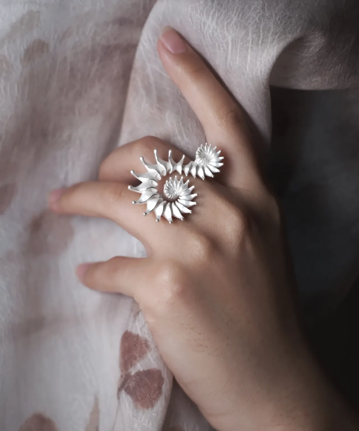 A close-up image of a hand adorned with the Spiral Ring, crafted from S925 sterling silver. The ring showcases an intricate sculptural design with elegant spiral patterns. The hand delicately holds a textured fabric, enhancing the ring’s artistic appeal.