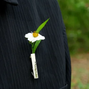 A close-up of a handmade 925 sterling silver brooch featuring a delicate white flower with a yellow center and vibrant green leaves, elegantly secured by a silver clip against a dark textured fabric background.