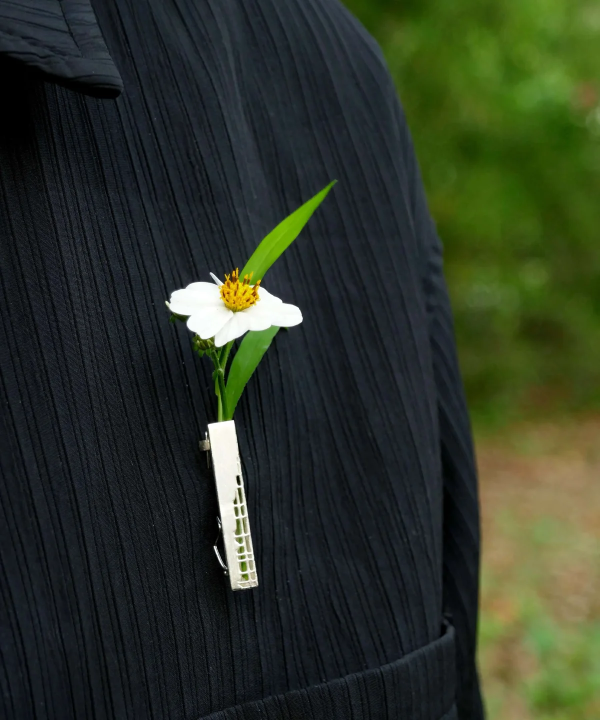 A close-up of a handmade 925 sterling silver brooch featuring a delicate white flower with a yellow center and vibrant green leaves, elegantly secured by a silver clip against a dark textured fabric background.