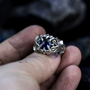 A close-up image of a hand holding the Vongola Ring Of Hell, a handmade sterling silver ring featuring a natural blue spinel gemstone. The ring's intricate design is inspired by Mukuro Rokudo from the anime Katekyo Hitman Reborn.