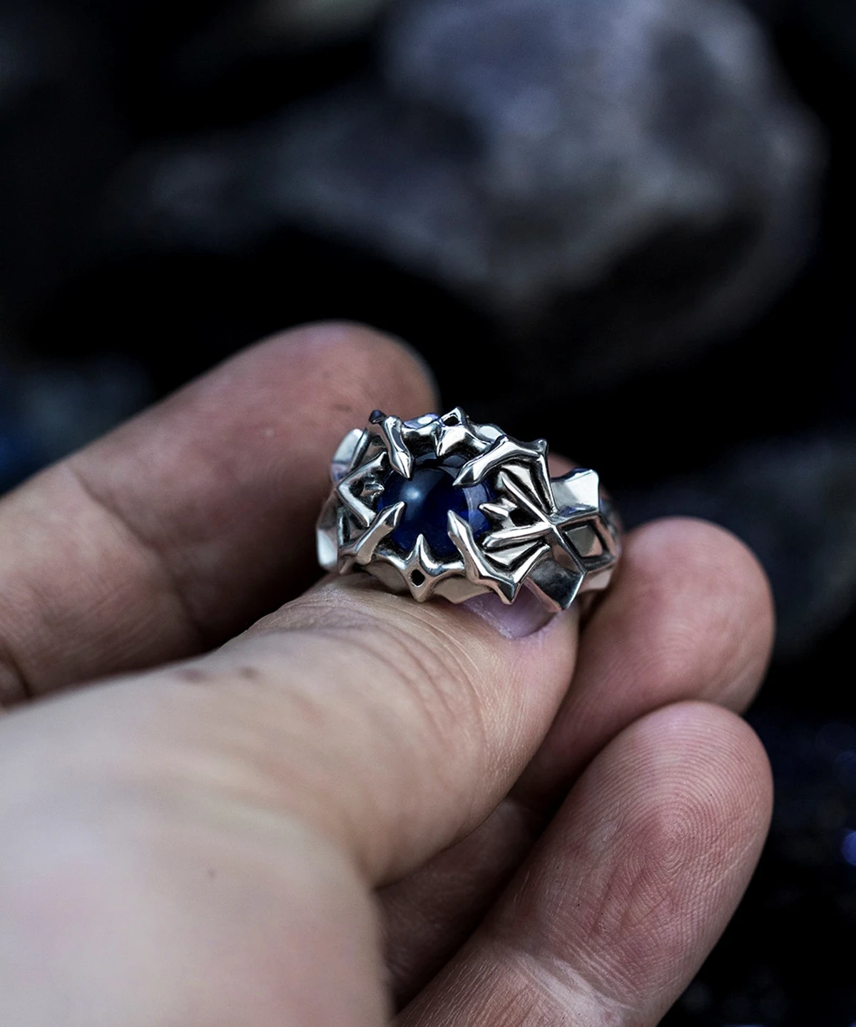 A close-up image of a hand holding the Vongola Ring Of Hell, a handmade sterling silver ring featuring a natural blue spinel gemstone. The ring's intricate design is inspired by Mukuro Rokudo from the anime Katekyo Hitman Reborn.
