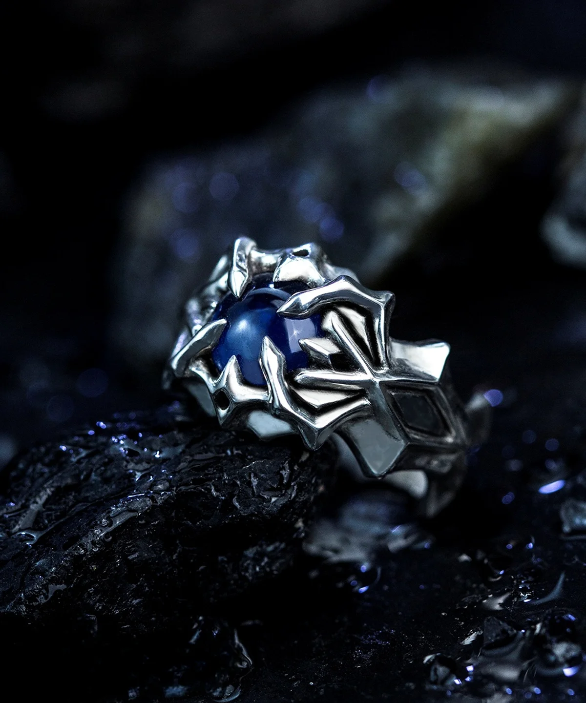 A close-up image of the Vongola Ring Of Hell, a handmade sterling silver ring featuring a natural blue spinel gemstone. Inspired by Mukuro Rokudo from Katekyo Hitman Reborn, the ring showcases intricate details and craftsmanship against a dark, rocky background.