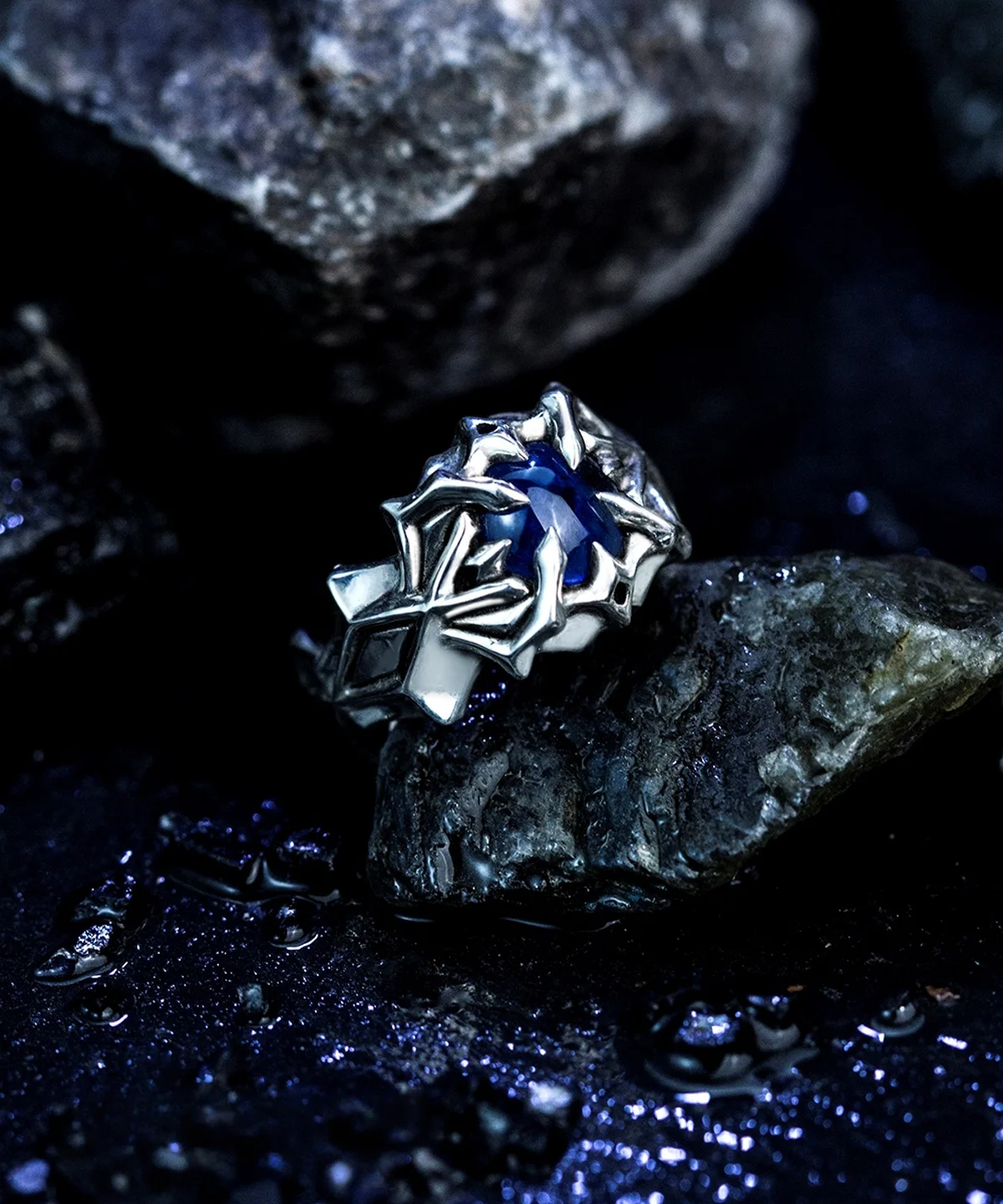 A close-up image of the Vongola Ring Of Hell, a handmade sterling silver ring featuring a natural blue spinel gemstone. Inspired by Mukuro Rokudo from Katekyo Hitman Reborn, the ring showcases intricate details and craftsmanship against a dark, rocky background.