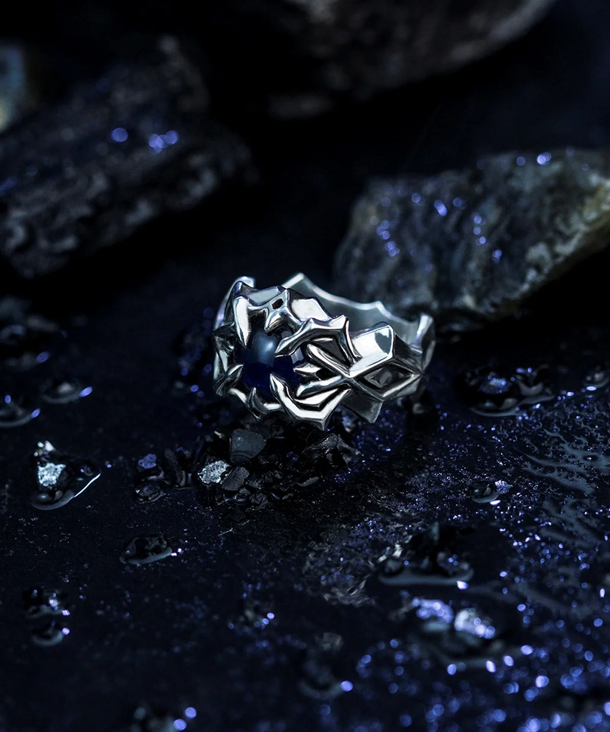 A close-up of the Vongola Ring Of Hell, a handcrafted S925 sterling silver ring featuring intricate detailing and a natural blue spinel gemstone. Inspired by Mukuro Rokudo from Katekyo Hitman Reborn, this anime jewelry piece is displayed on a dark, textured surface with scattered reflective elements, enhancing its allure.
