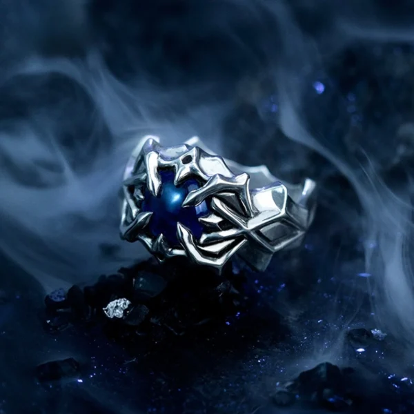 A close-up image of the Vongola Ring Of Hell, a handmade sterling silver ring featuring a natural blue spinel gemstone. The ring has intricate, claw-like designs inspired by Mukuro Rokudo from the anime Katekyo Hitman Reborn. The background is dark and smoky, enhancing the ring's mystical appearance.