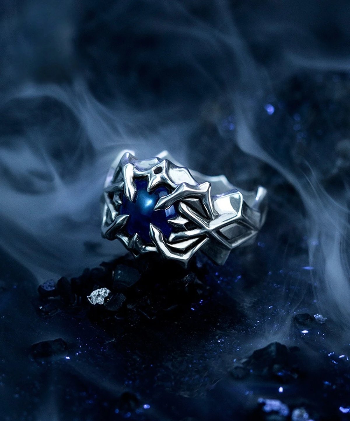 A close-up image of the Vongola Ring Of Hell, a handmade sterling silver ring featuring a natural blue spinel gemstone. The ring has intricate, claw-like designs inspired by Mukuro Rokudo from the anime Katekyo Hitman Reborn. The background is dark and smoky, enhancing the ring's mystical appearance.