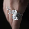 Home 54 Close-up of a Lazesoul Orchid Ring crafted in S925 sterling silver, featuring delicate orchid petals and an adjustable band; nature-inspired handmade jewelry styled on a woman's hand. Orchid Ring design highlights floral elegance and artisanal detail.