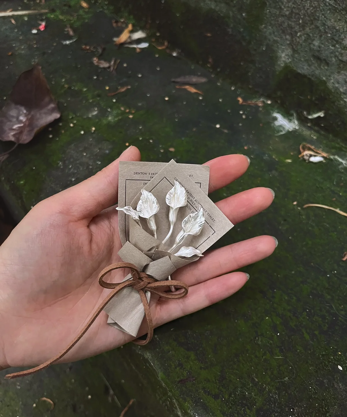 Hand holding a delicate 925 sterling silver mini Calla Lily Bouquet, wrapped in rustic brown paper with string bow, Lazesoul Handmade Art Gifts, nature-inspired jewelry perfect for meaningful art gifts.