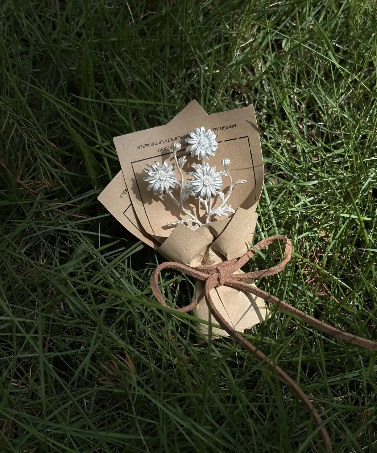 Handmade 925 sterling silver mini daisy bouquet brooch, presented as an elegant art gift. White floral design wrapped in rustic brown paper on green grass, perfect for nature-inspired jewelry lovers and Daisy Bouquet collectors.