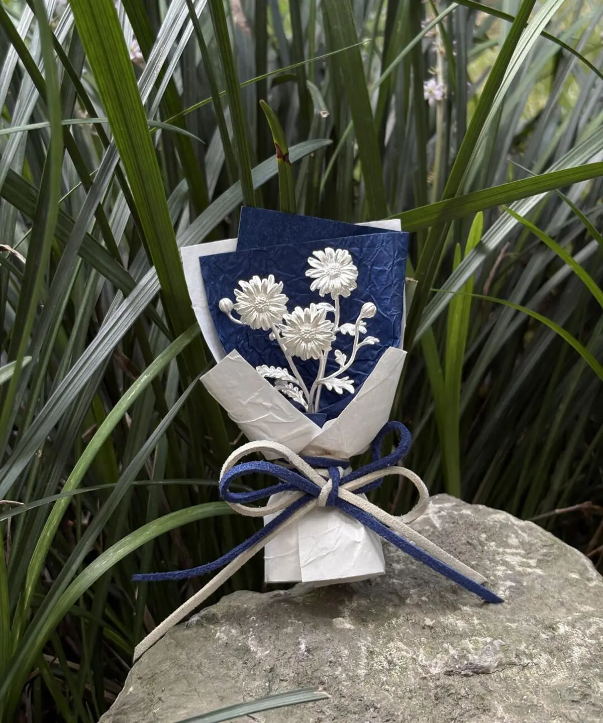 Handmade 925 sterling silver mini daisy bouquet displayed on a rock with green grass backdrop. Elegant white daisies on deep blue paper, wrapped with dual‑color bow. Perfect daisy bouquet for unique handmade art gifts by Lazesoul.