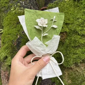 Hand holding a delicate paper-crafted Gardenia Bouquet against a mossy tree trunk, inspired by Lazesoul Handmade Art Gifts. Features 925 sterling silver mini gardenia design, perfect for nature lovers and elegant art gifts.