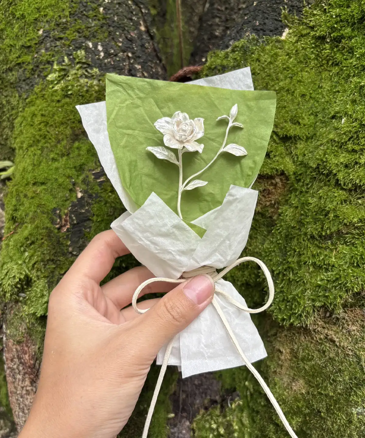 Hand holding a delicate paper-crafted Gardenia Bouquet against a mossy tree trunk, inspired by Lazesoul Handmade Art Gifts. Features 925 sterling silver mini gardenia design, perfect for nature lovers and elegant art gifts.