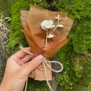 Hand holding a rustic mini Gardenia Bouquet wrapped in brown paper with twine, set against mossy tree bark. Lazesoul Handmade Art Gifts – 925 Sterling Silver Mini Gardenia Bouquet, nature-inspired elegance.