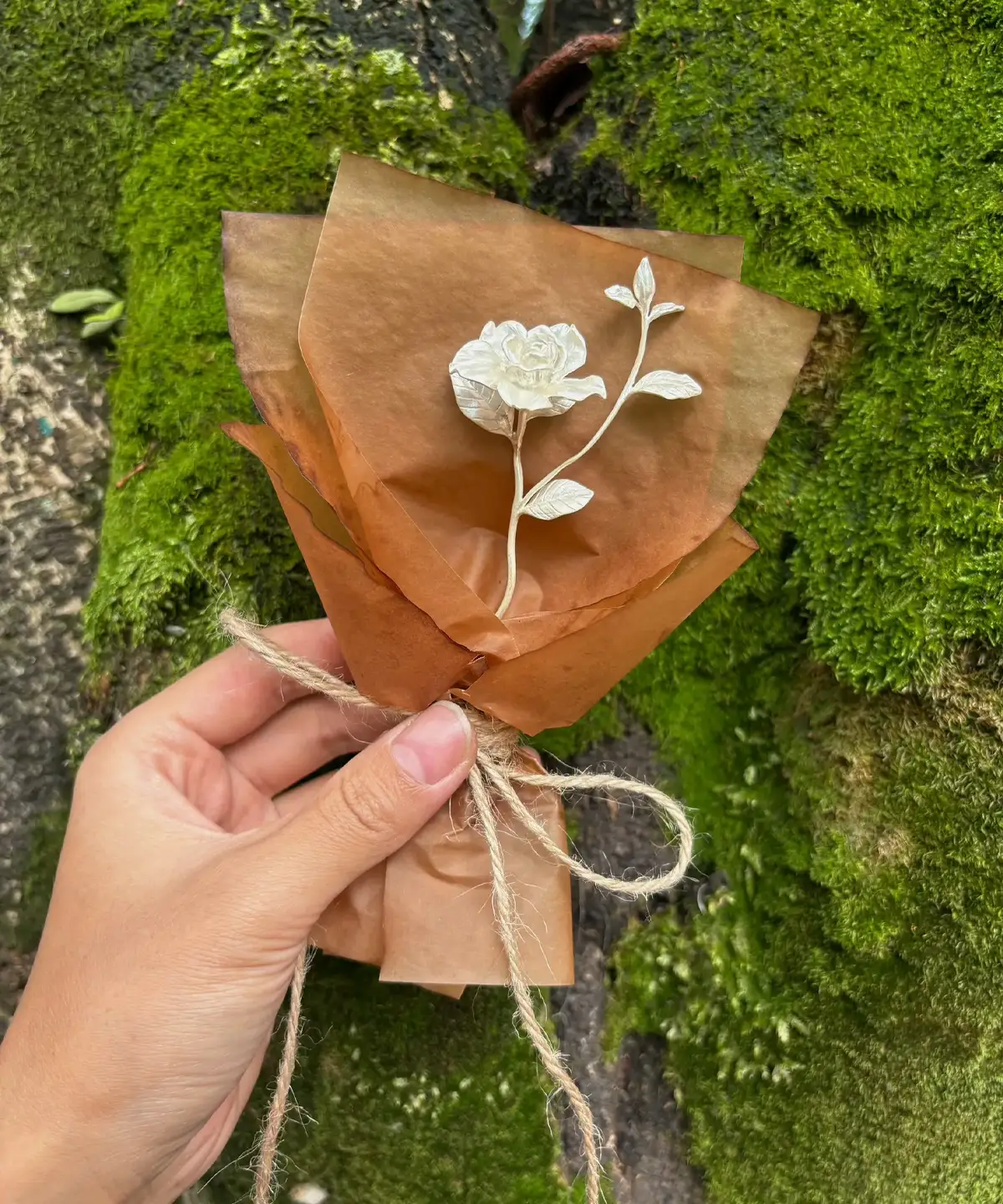 Hand holding a rustic mini Gardenia Bouquet wrapped in brown paper with twine, set against mossy tree bark. Lazesoul Handmade Art Gifts – 925 Sterling Silver Mini Gardenia Bouquet, nature-inspired elegance.