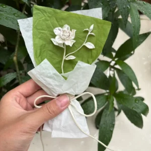 Hand holding a delicate 925 sterling silver mini gardenia bouquet, wrapped in white tissue with bow, against fresh green leaves. Handmade art gift by Lazesoul, perfect for nature‑inspired jewelry lovers.