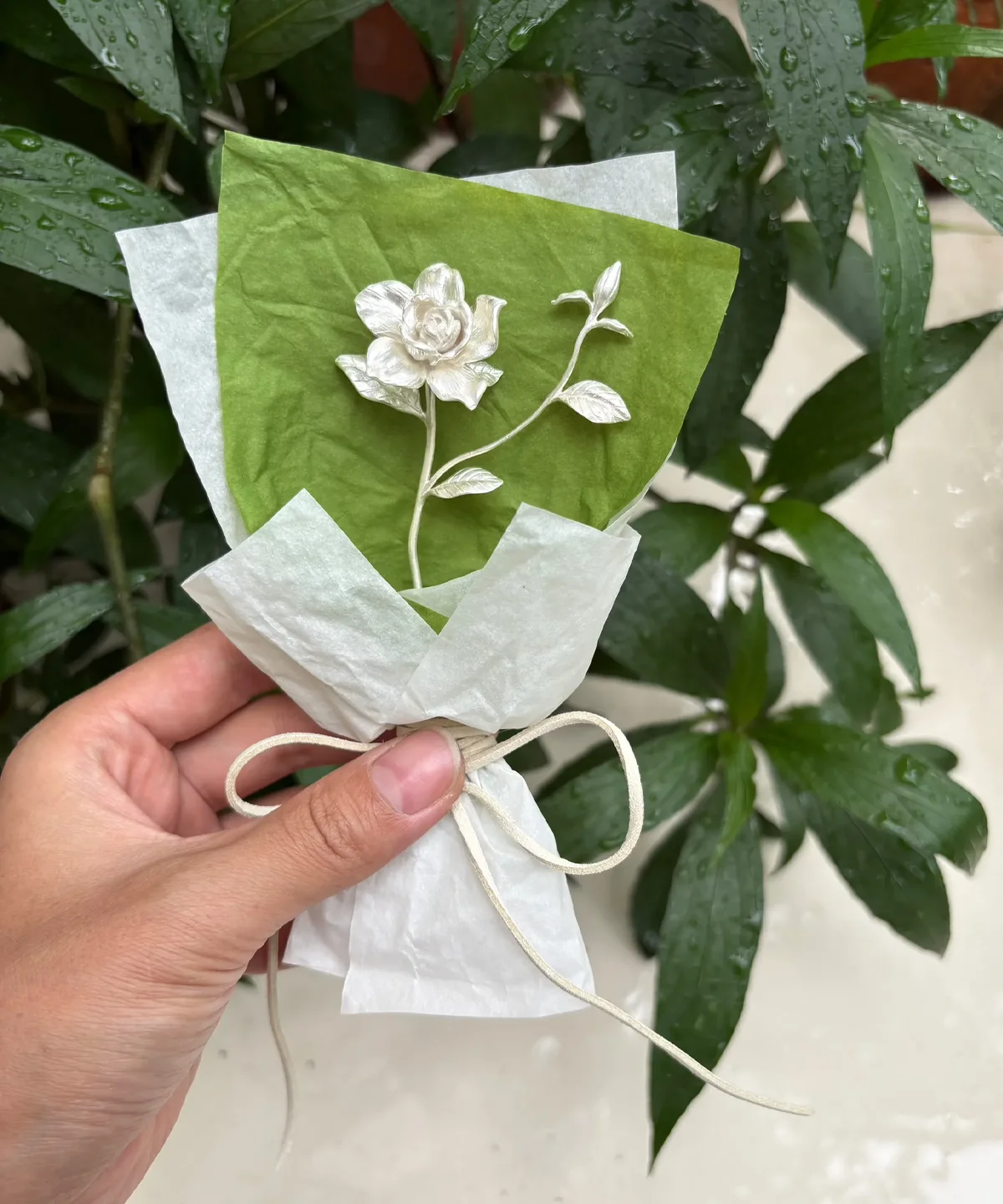 Hand holding a delicate 925 sterling silver mini gardenia bouquet, wrapped in white tissue with bow, against fresh green leaves. Handmade art gift by Lazesoul, perfect for nature‑inspired jewelry lovers.