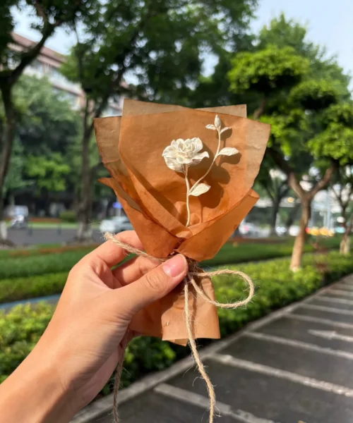 A hand holds a rustic mini Gardenia Bouquet wrapped in brown paper with twine, featuring a single white bloom. Perfect as handmade art gifts, inspired by Lazesoul’s 925 sterling silver jewelry design and nature’s timeless elegance.