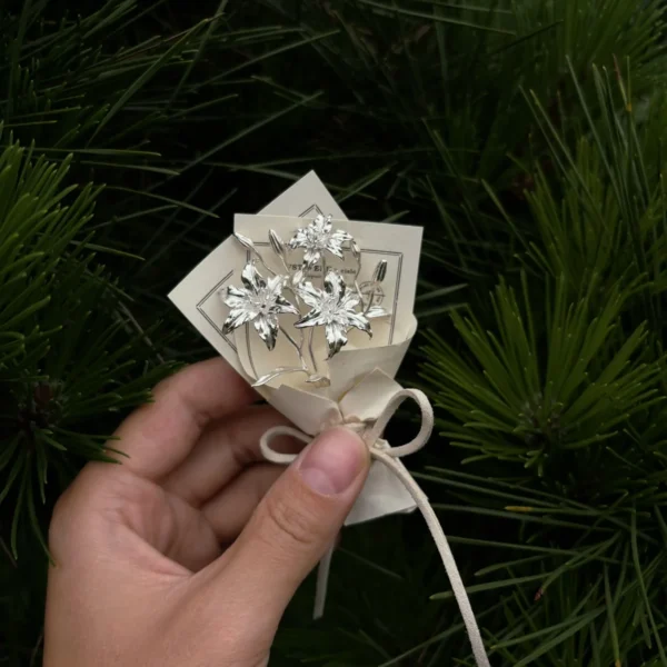Hand holding a 925 sterling silver mini lily bouquet wrapped in cream paper with string bow, set against lush green pine background. Handmade art gifts by Lazesoul, perfect for elegant décor, keepsakes, and unique Lily Bouquet lovers.