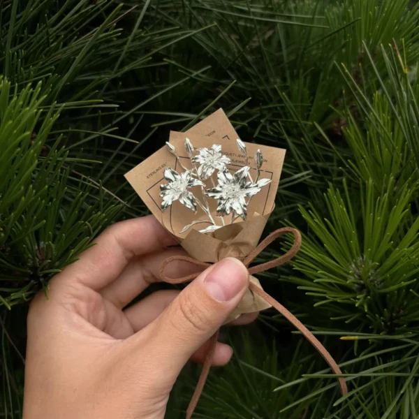 Hand holding a delicate mini lily bouquet wrapped in brown paper with two small cards, set against lush green pine needles. Perfect for Lazesoul handmade art gifts, 925 sterling silver Lily Bouquet jewelry and keepsakes.